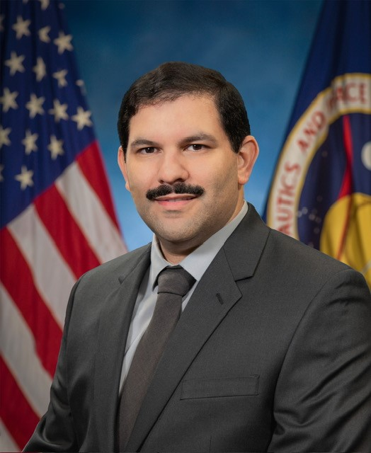 A person in a dark grey suit in front of a blue background 